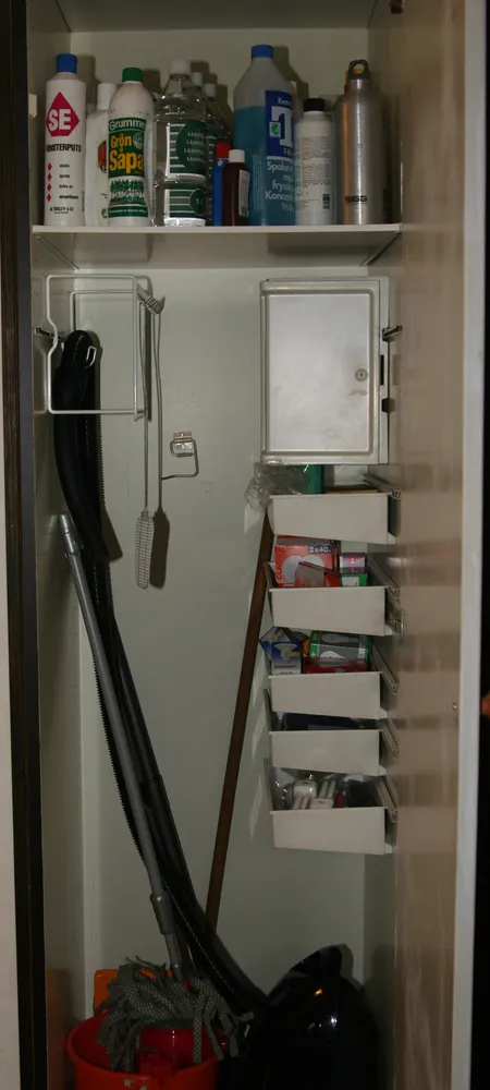 Narrow broom cupboard with cleaning bottles on a top shelf, a vacuum hose and a mop hung on wall hooks, and a column of pull-out trays on the side wall. Photo: Dnor / Wikimedia Commons, public domain